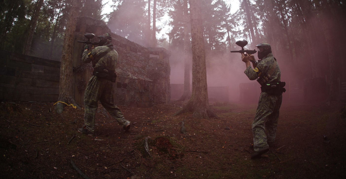 Two men wearing camouflage gear are seen shooting paintballs in a forest, highlighting an exciting outdoor sport